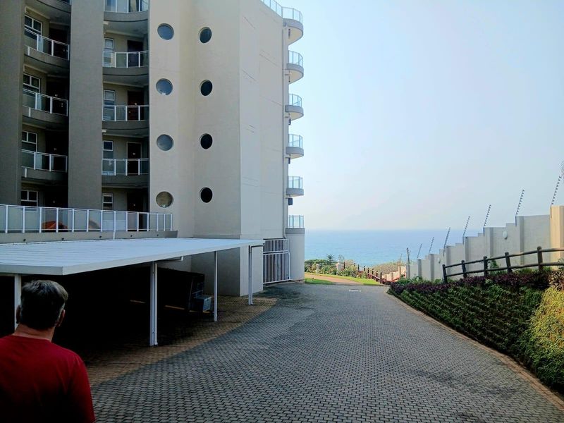 Exterior view of an apartment complex featuring a paved driveway, covered parking, and ocean views with secure perimeter fencing.