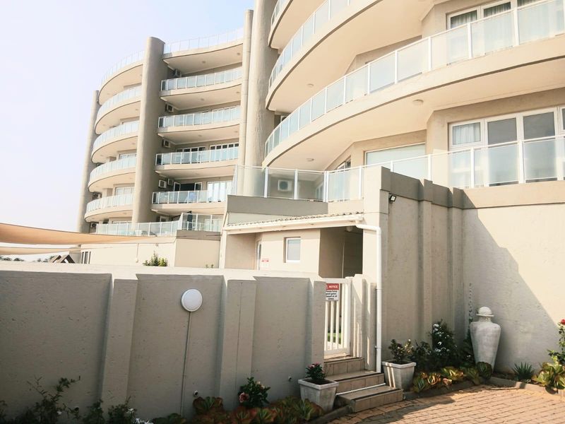 Exterior view of a modern multi-story apartment building featuring curved balconies with glass balustrades and a paved entrance area.