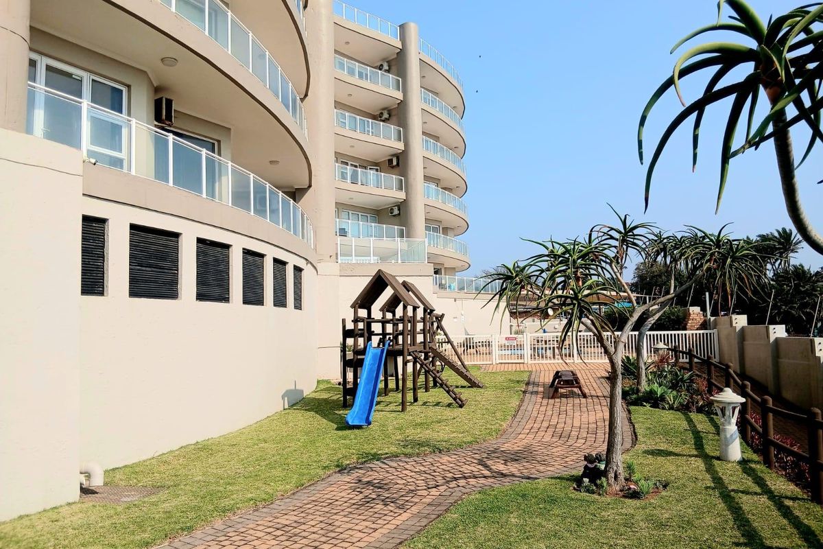 A well-maintained rear garden area of an apartment complex featuring a paved walkway, lawn, and glass-balustraded balconies on the building facade.