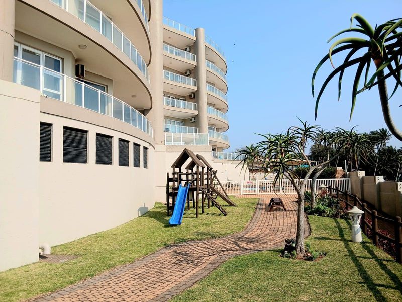 A well-maintained rear garden area of an apartment complex featuring a paved walkway, lawn, and glass-balustraded balconies on the building facade.