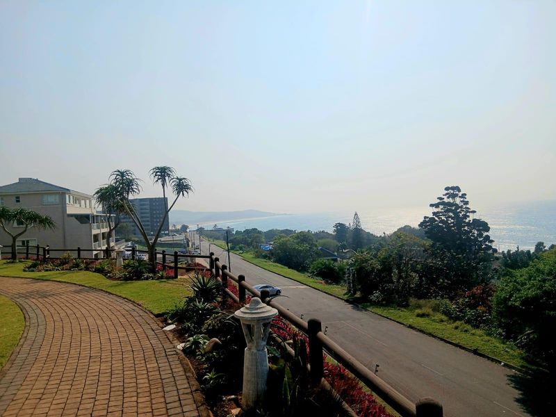 Elevated coastal view overlooking the road, surrounding greenery, and the ocean horizon, featuring a paved walkway and landscaped garden area.