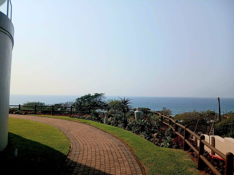 A scenic outdoor view featuring a curved paved walkway, landscaped garden beds, and timber fencing overlooking the ocean.