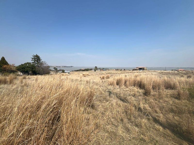 A wide-angle view of a vacant residential plot featuring dry grassland with distant water views and surrounding neighborhood development.