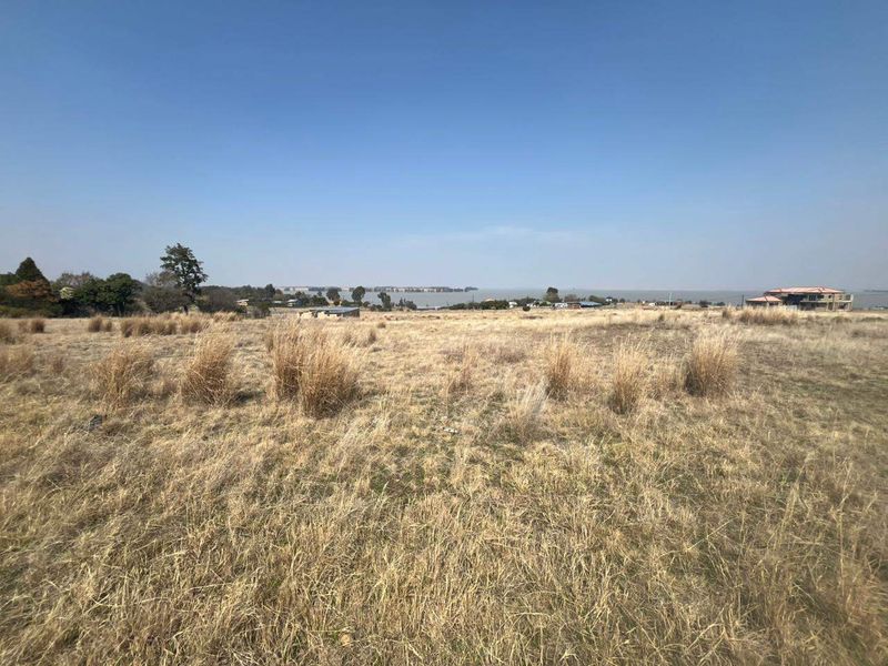 A wide-angle view of a vacant residential plot featuring natural dry grass landscape with distant water views.