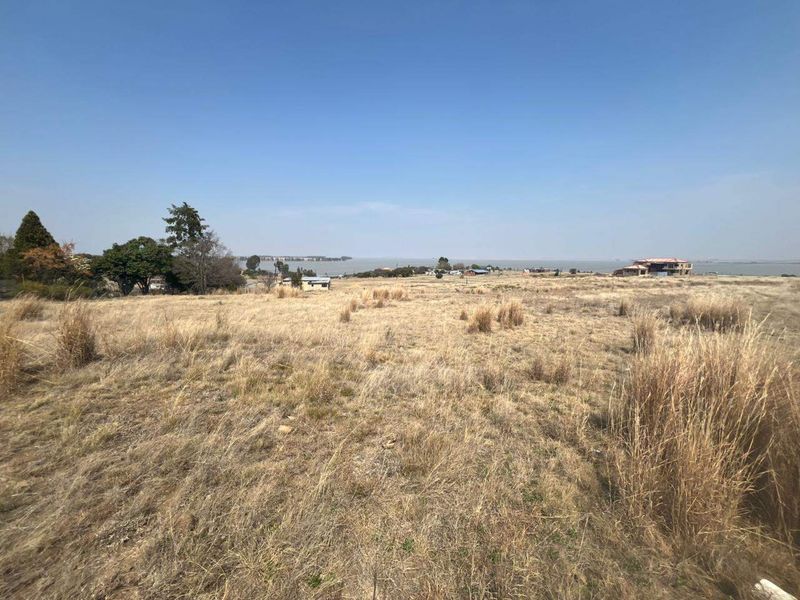 A wide-angle view of a vacant residential plot featuring dry grass and a distant water landscape.