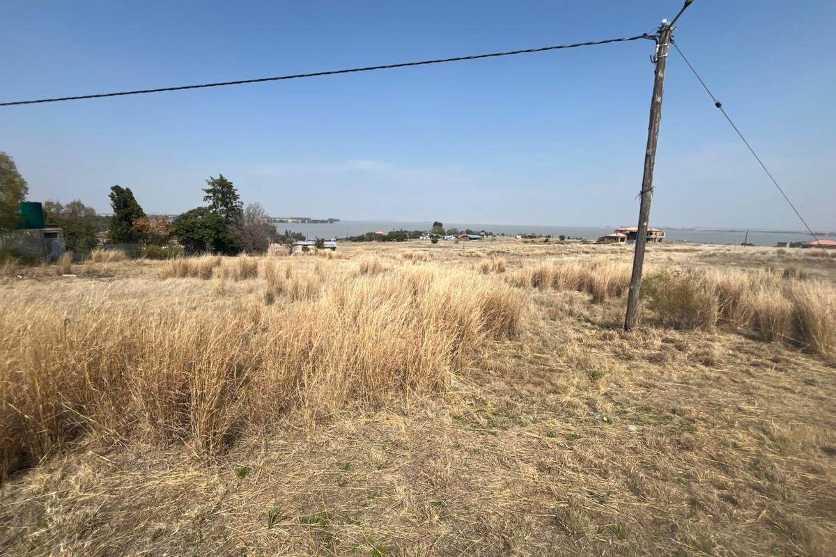 A wide-angle view of a vacant residential plot featuring dry grass, a utility pole, and distant water views.