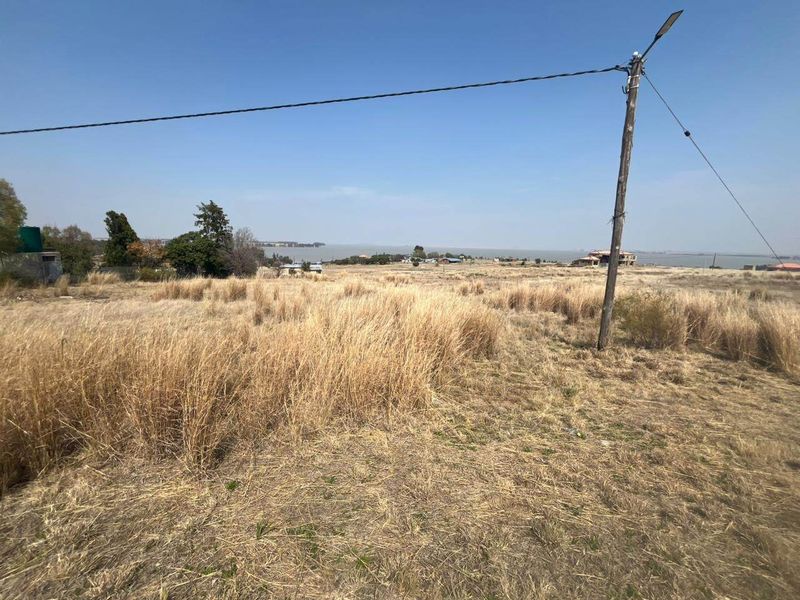 A wide-angle view of a vacant residential plot featuring dry grass, a utility pole, and distant water views.