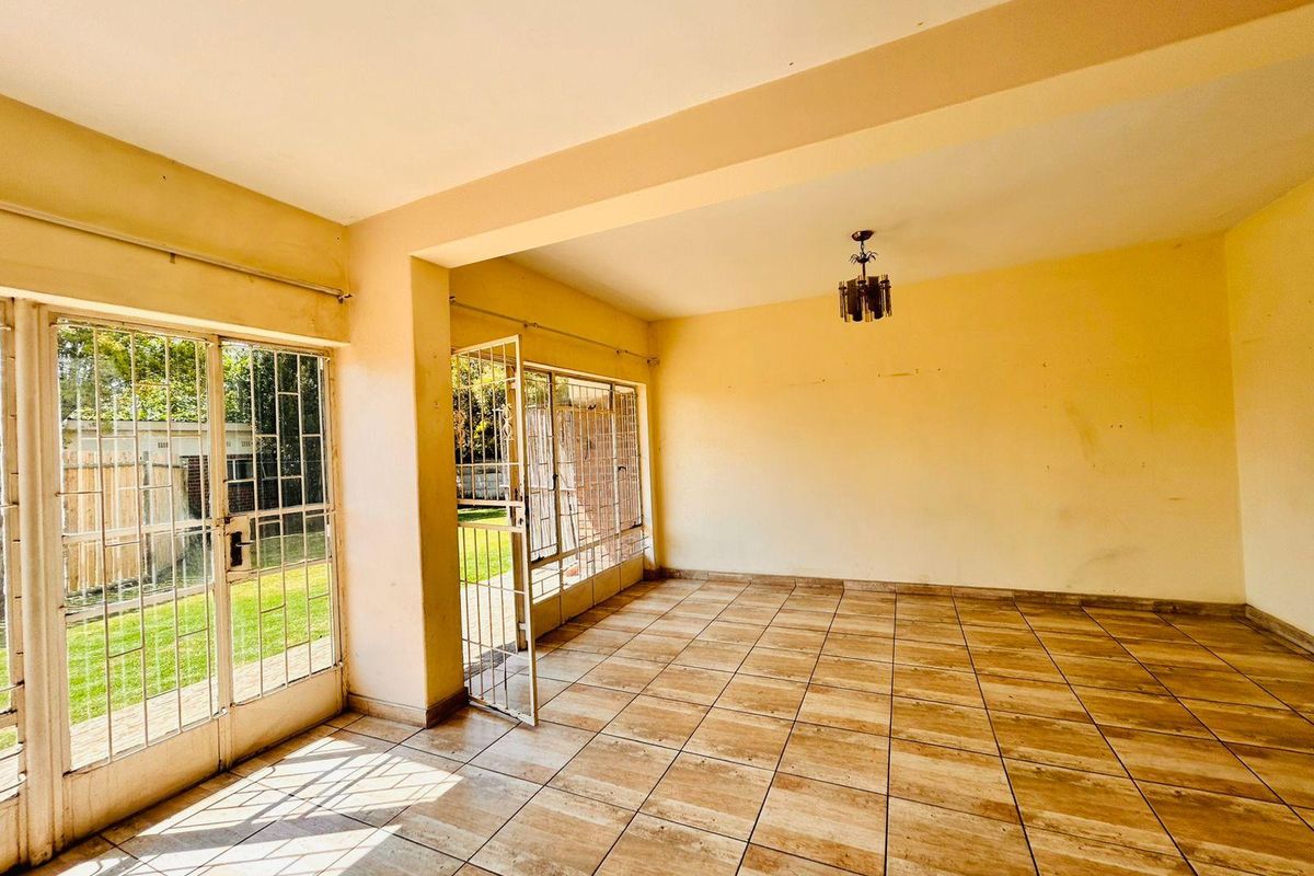 An empty, sunlit living room featuring tiled flooring and large glass sliding doors with security gates, providing direct access to the garden area.