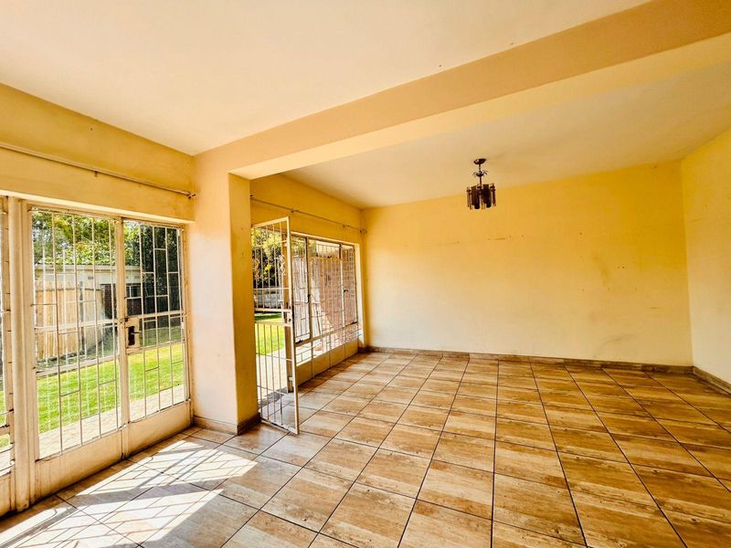 An empty, sunlit living room featuring tiled flooring and large glass sliding doors with security gates, providing direct access to the garden area.
