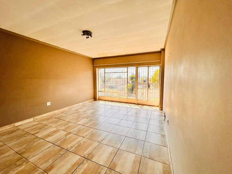 An empty living room featuring tiled flooring, neutral walls, and large windows secured with metal security bars and a gate.