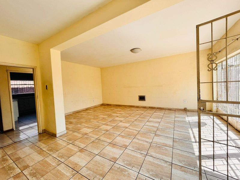 An empty, open-plan living area featuring tiled flooring, a wall-mounted heater, and a security gate leading to the exterior.