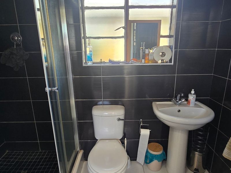 Compact bathroom featuring a shower enclosure, pedestal basin, and toilet, finished with dark wall tiles and a metal-framed window.