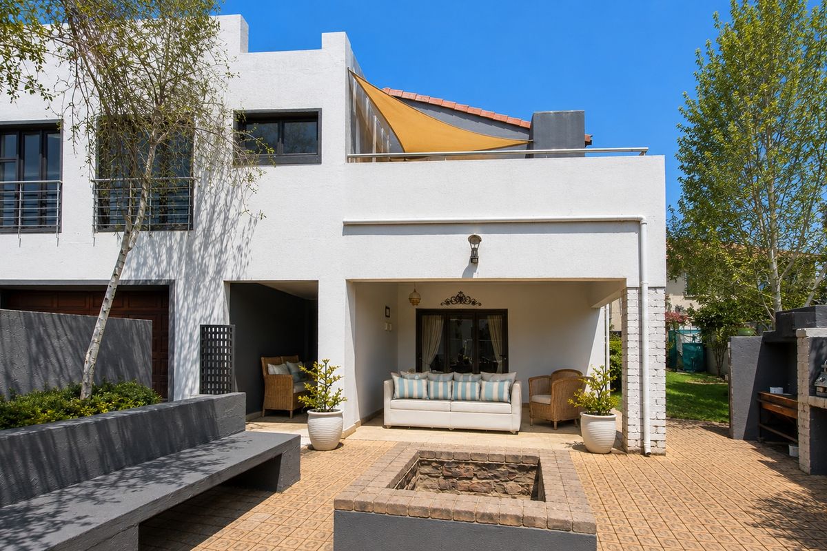 Spacious rear exterior featuring a covered patio, built-in braai, fire pit, and a paved courtyard area with an upper-level balcony.