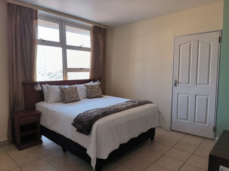 A well-lit bedroom featuring tiled flooring, a built-in headboard, and large windows allowing for natural light.