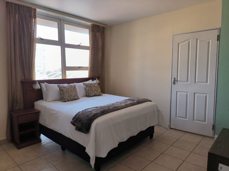 A functional bedroom featuring tiled flooring, a built-in headboard, and a large window providing natural light.