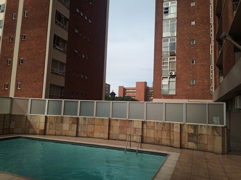 Communal swimming pool area featuring stone-clad walls, a frosted glass privacy screen, and tiled decking situated between apartment blocks.