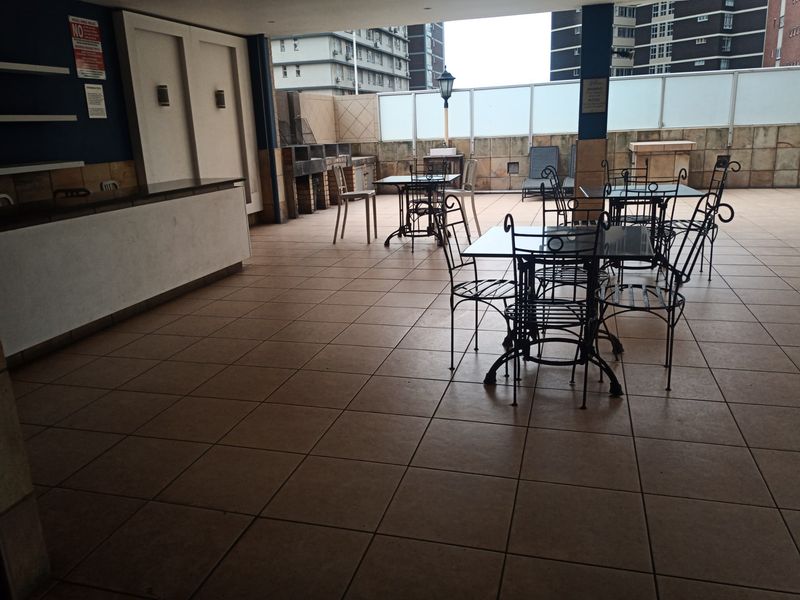 A covered outdoor patio area featuring tiled flooring, a built-in braai, and glass balustrades, suitable for communal or private outdoor entertainment.