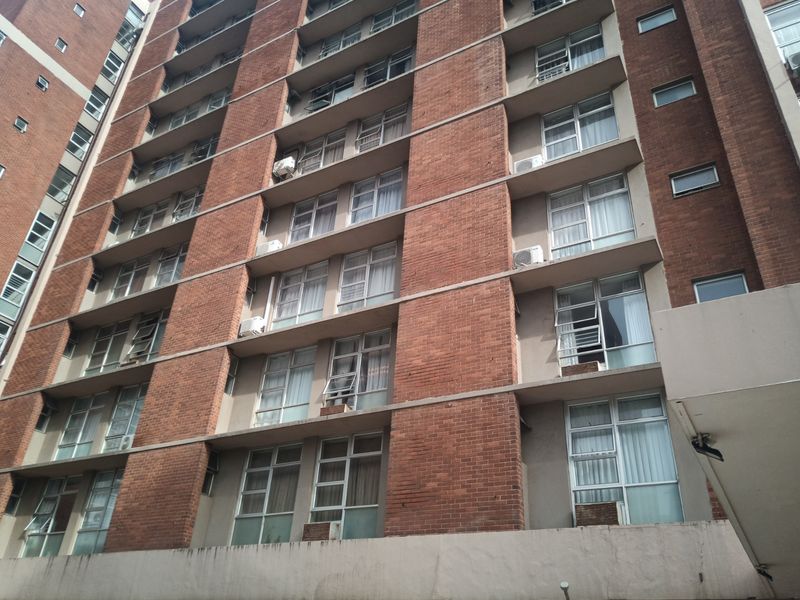 A low-angle view of a multi-story apartment building facade featuring red brick construction, aluminum-framed windows, and integrated air conditioning units.