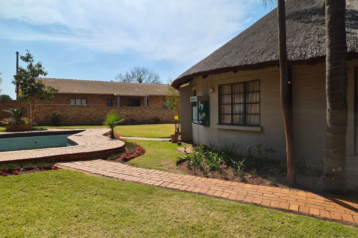 A well-maintained rear garden featuring a swimming pool, brick pathways, and a traditional thatched-roof structure set against a brick boundary wall.