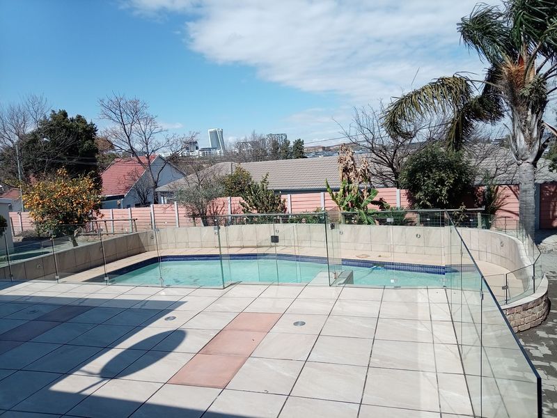 Outdoor pool area featuring a tiled patio, glass safety balustrade, and a swimming pool with a view of the surrounding neighborhood.