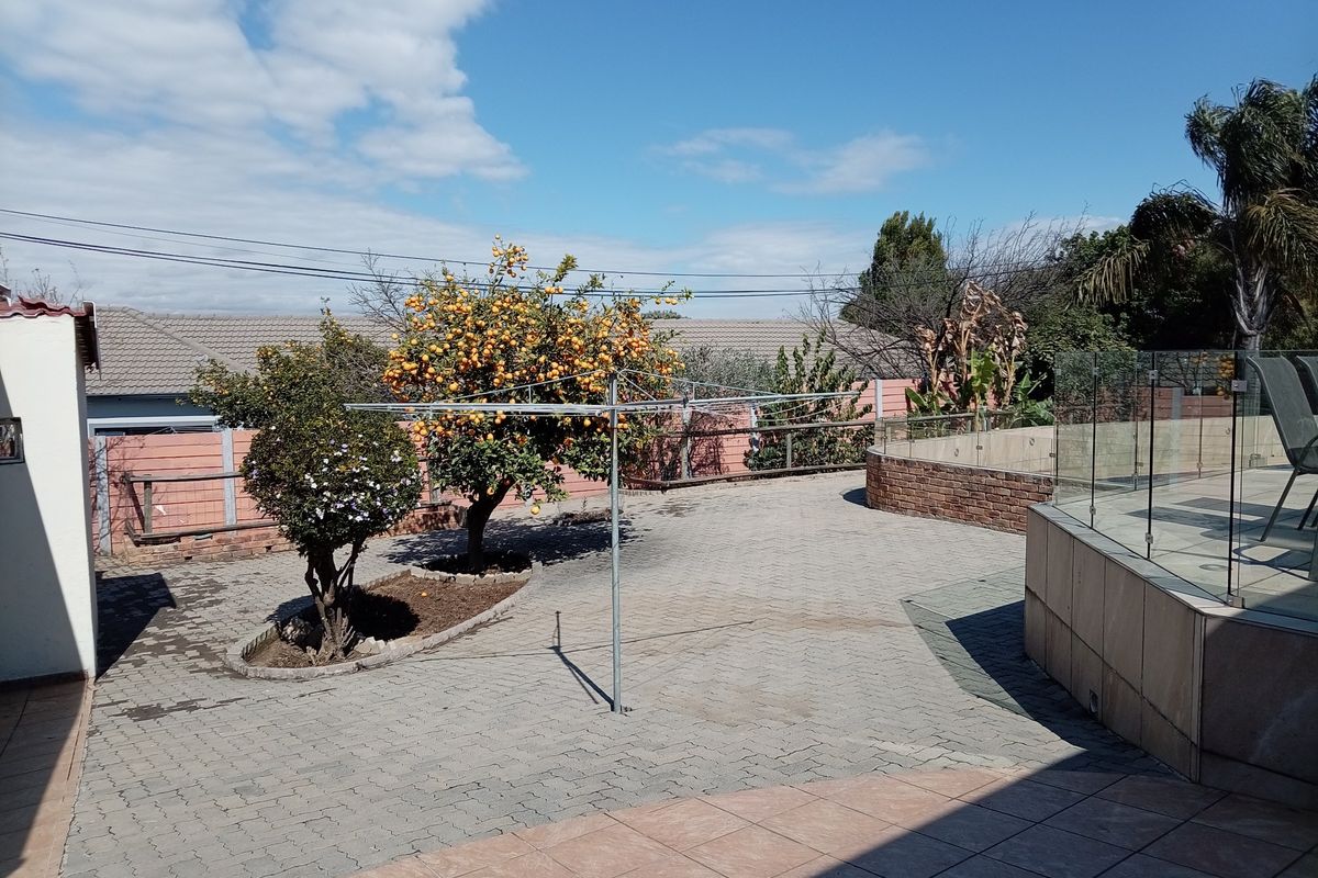 A spacious rear paved patio area featuring a glass balustrade, brick retaining wall, and established fruit trees within a secure perimeter.