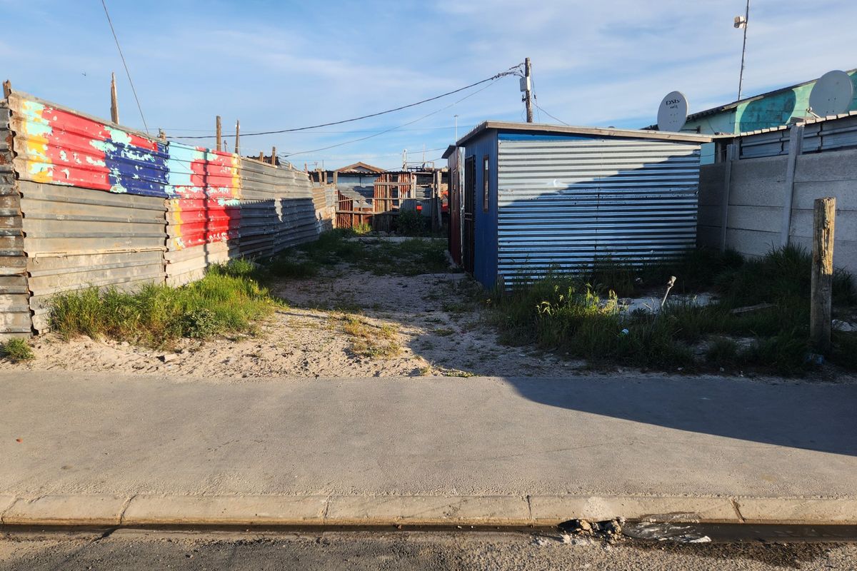 Vacant residential plot featuring corrugated iron boundary fencing and a small metal structure, accessible from a paved street.