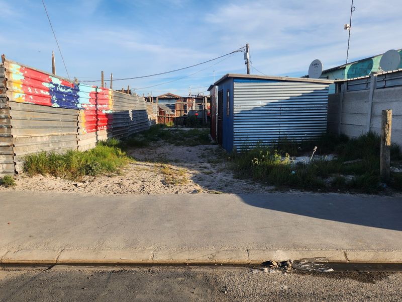 Vacant residential plot featuring corrugated iron boundary fencing and a small metal structure, accessible from a paved street.