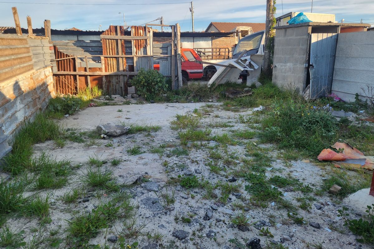 A vacant residential plot featuring a concrete slab, corrugated metal fencing, and a small utility structure, currently requiring significant clearing and site preparation.