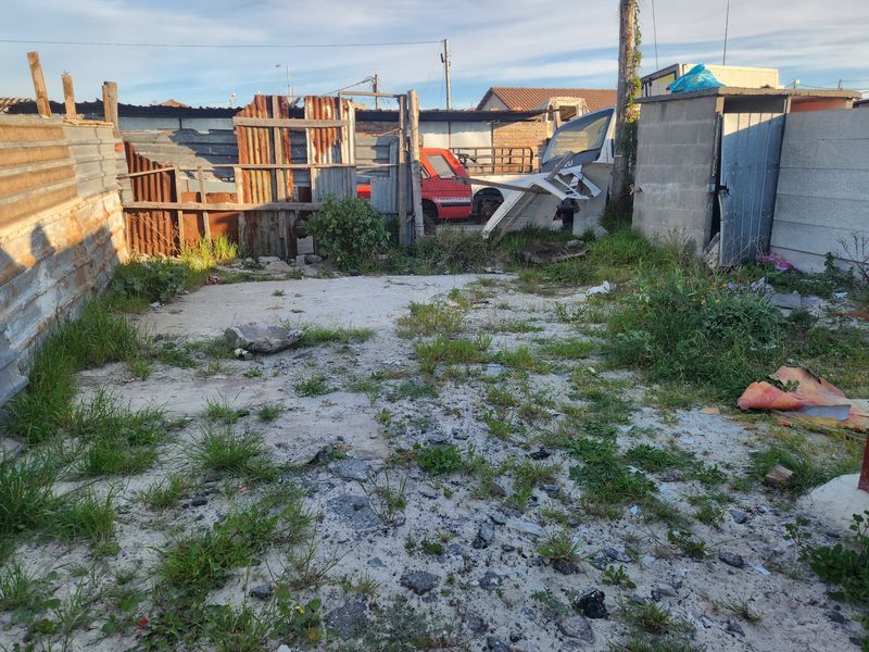 A vacant residential plot featuring a concrete slab, corrugated metal fencing, and a small utility structure, currently requiring significant clearing and site preparation.