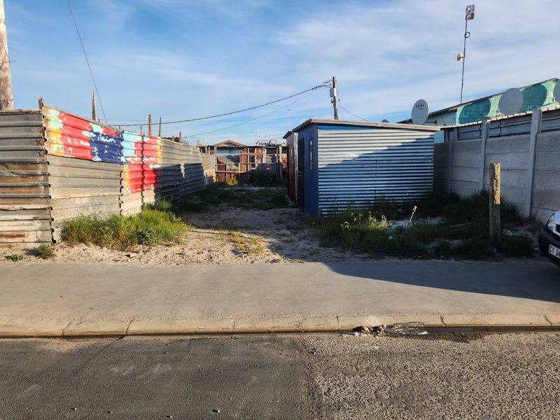 Vacant residential plot featuring corrugated metal fencing, a small utility shed, and direct street access via a paved sidewalk.