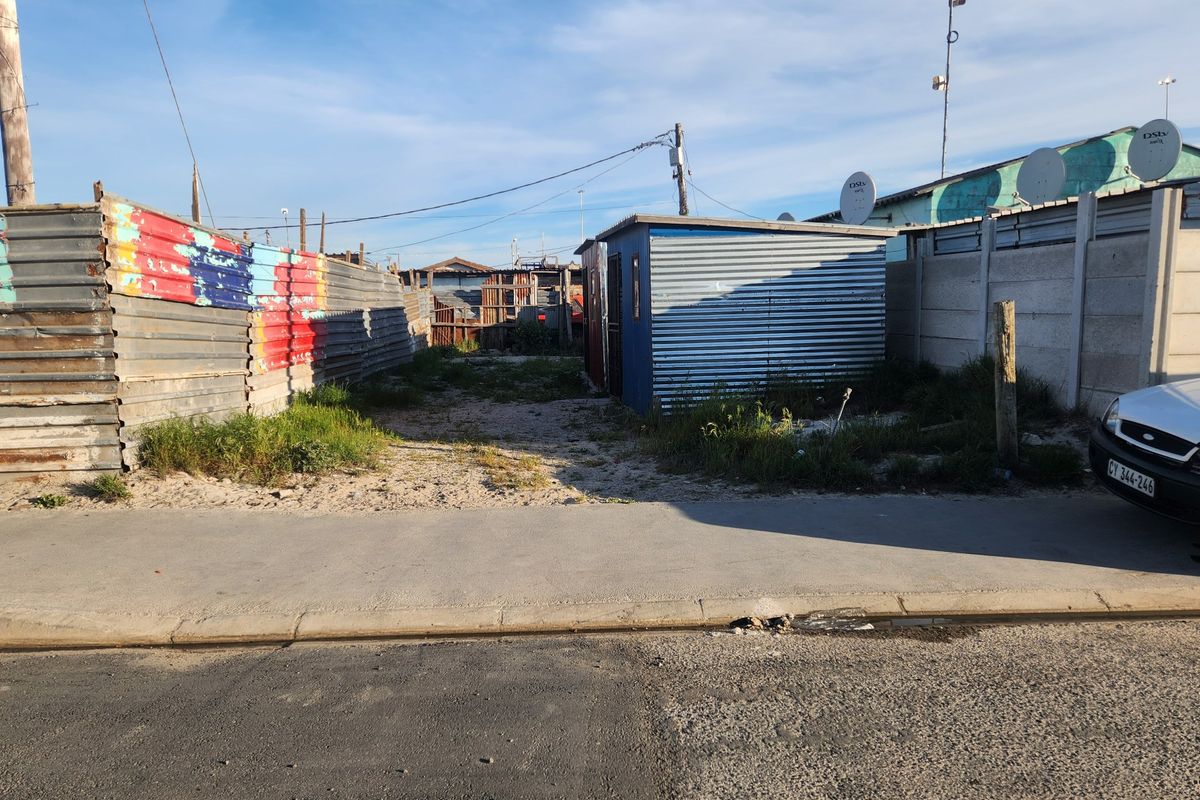 A vacant residential plot featuring corrugated metal fencing, a small utility shed, and direct street access from the paved sidewalk.