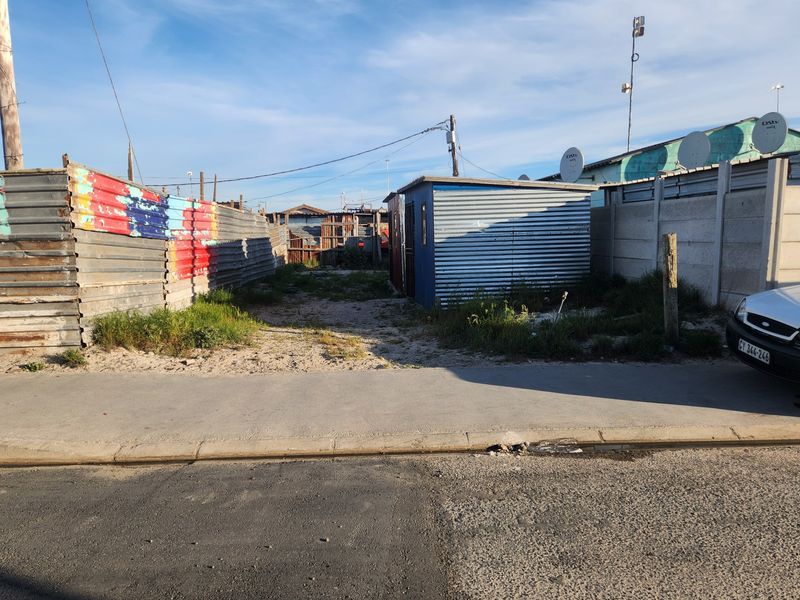 A vacant residential plot featuring corrugated metal fencing, a small utility shed, and direct street access from the paved sidewalk.