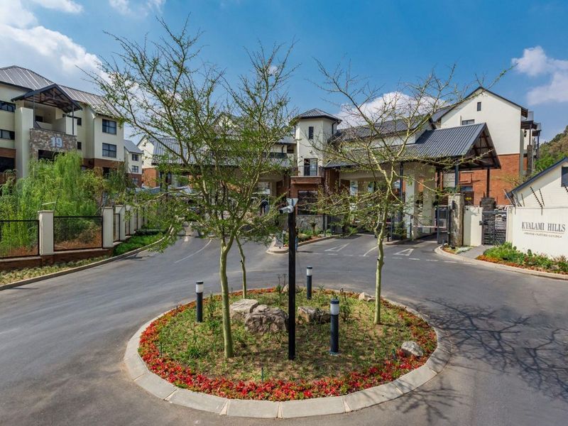 The main entrance and security gatehouse of the Kyalami Hills apartment complex, featuring a paved driveway, landscaped traffic island, and secure perimeter fencing.