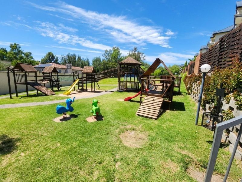 A well-maintained communal garden area featuring a wooden playground structure, lawn, and perimeter fencing within the apartment complex.