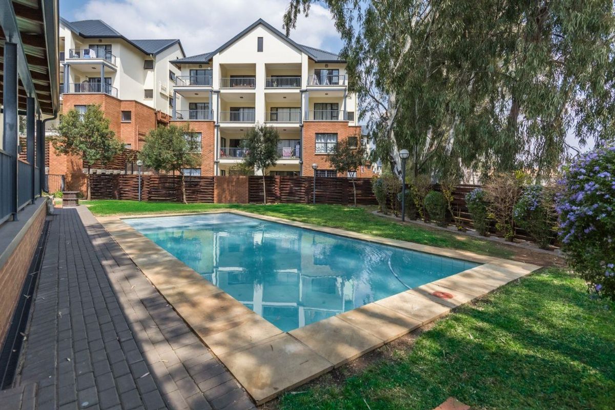 A well-maintained communal swimming pool area featuring paved surrounds, landscaped garden beds, and timber fencing, set against a multi-story apartment building.