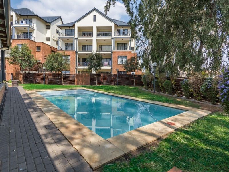 A well-maintained communal swimming pool area featuring paved surrounds, landscaped garden beds, and timber fencing, set against a multi-story apartment building.