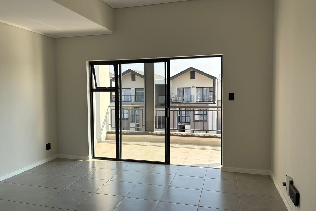 A bright, empty living area featuring neutral tiled flooring and a large sliding glass door providing direct access to a private balcony.