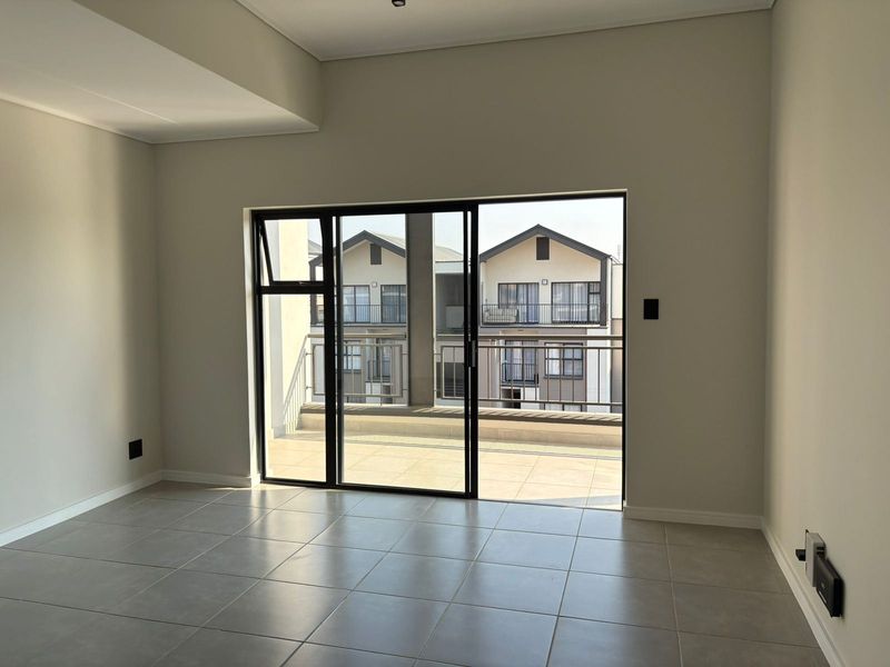 A bright, empty living area featuring neutral tiled flooring and a large sliding glass door providing direct access to a private balcony.