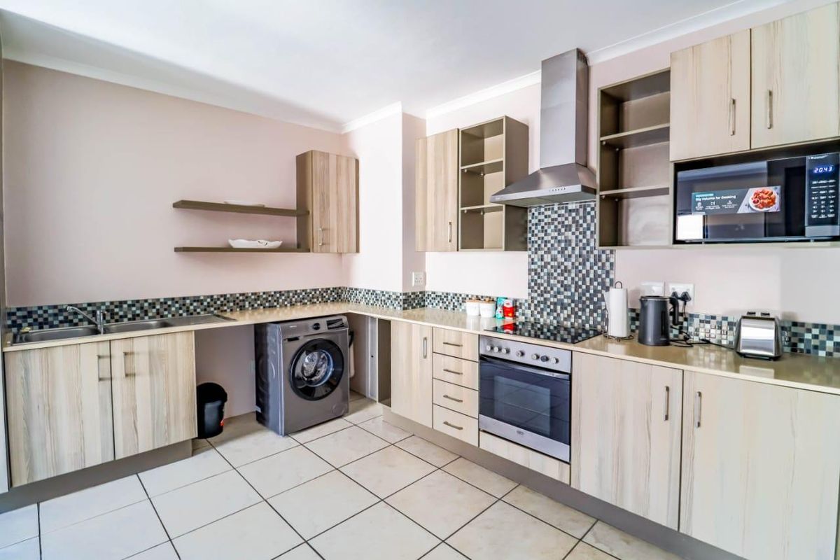 A functional kitchen featuring light wood-finish cabinetry, mosaic tile backsplash, integrated stainless steel appliances, and ceramic floor tiling.