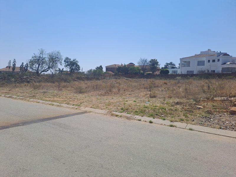 A vacant residential plot of land viewed from the adjacent paved street, showing natural vegetation and surrounding residential development.