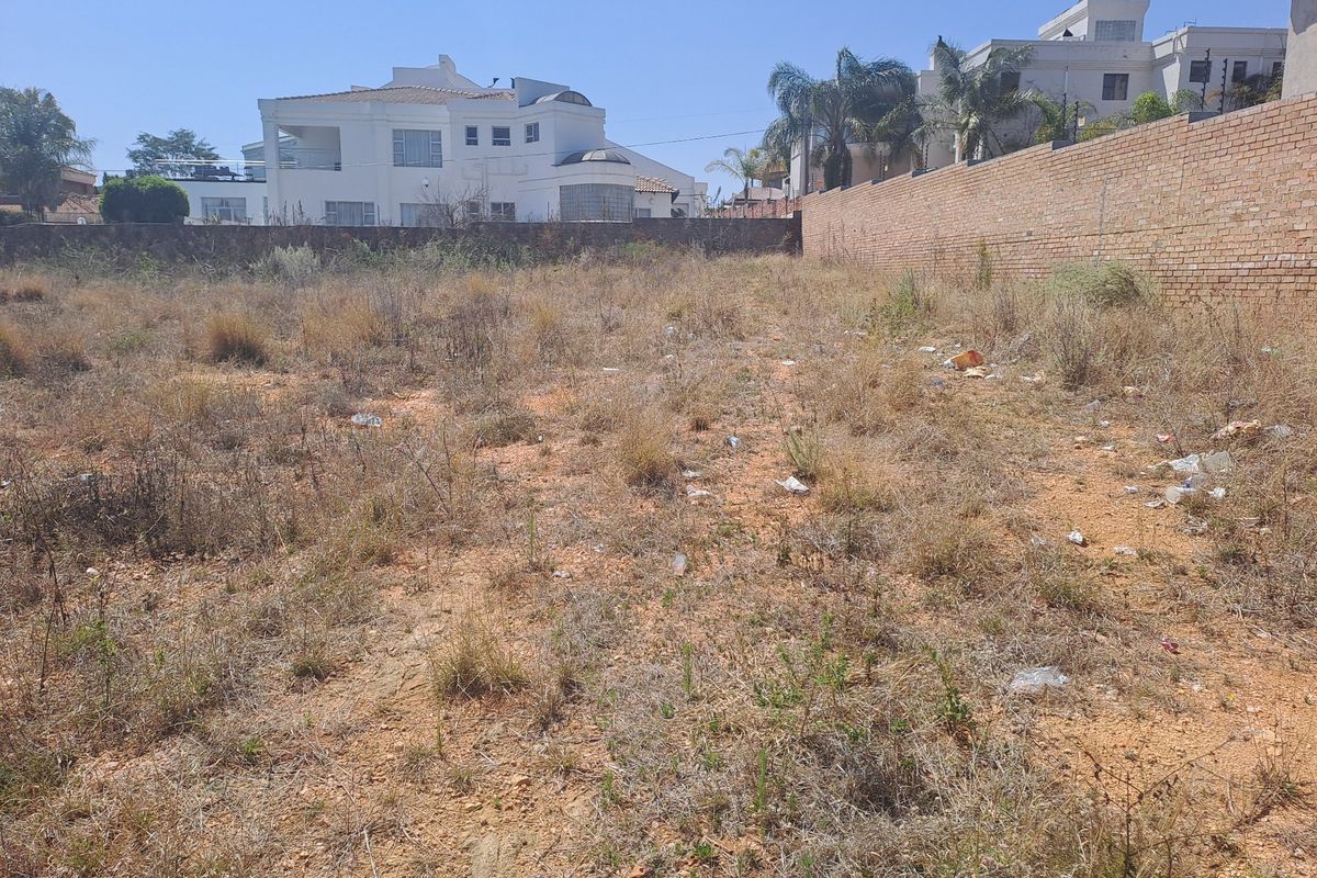 A vacant residential plot featuring dry, overgrown vegetation and a perimeter brick wall, situated in an established neighborhood.
