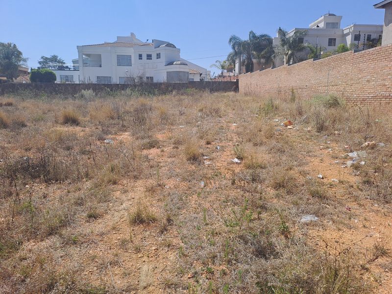 A vacant residential plot featuring dry, overgrown vegetation and a perimeter brick wall, situated in an established neighborhood.