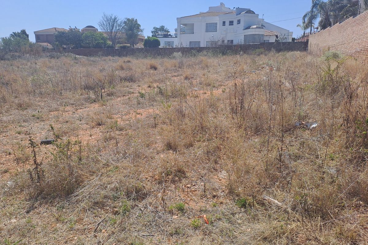 A vacant residential plot featuring dry, overgrown vegetation and a perimeter brick wall, situated in a developed neighborhood.