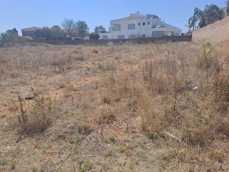 A vacant residential plot featuring dry, overgrown vegetation and a perimeter brick wall, situated in a developed neighborhood.
