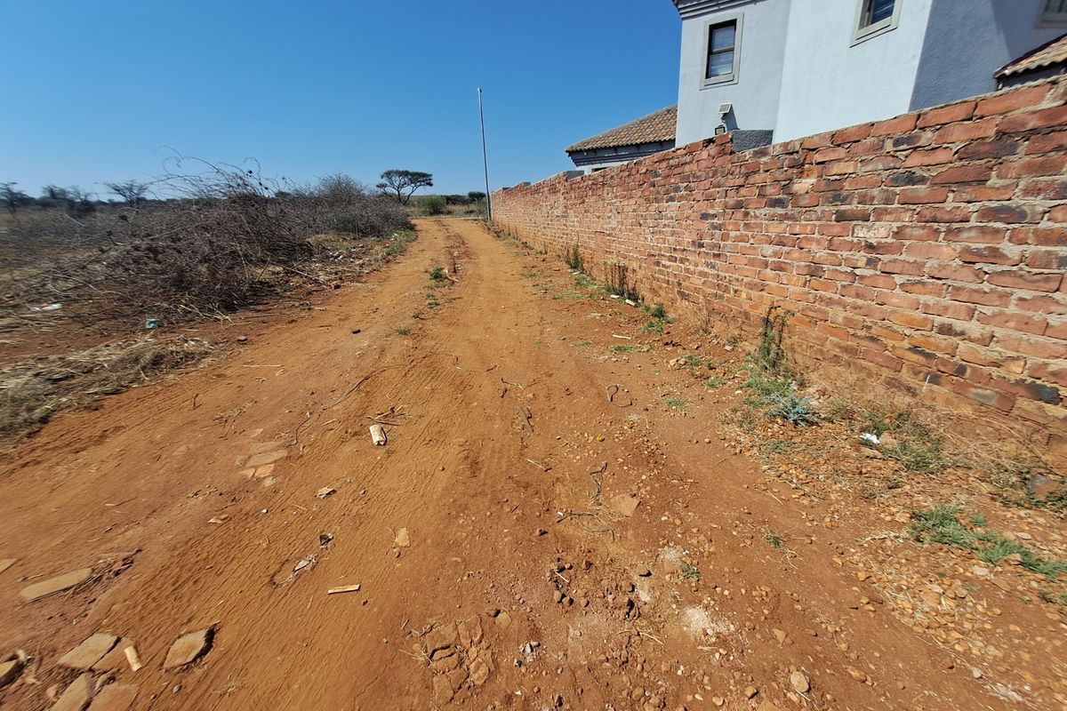 A view of an unpaved access road running alongside a residential brick boundary wall on a vacant land plot.