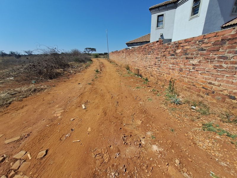 A view of an unpaved access road running alongside a residential brick boundary wall on a vacant land plot.
