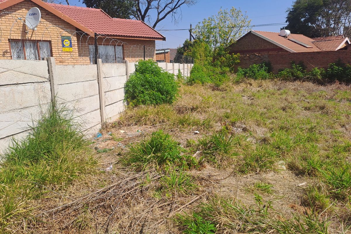 A vacant residential plot featuring a precast concrete boundary wall with security razor wire, adjacent to existing brick residential properties.