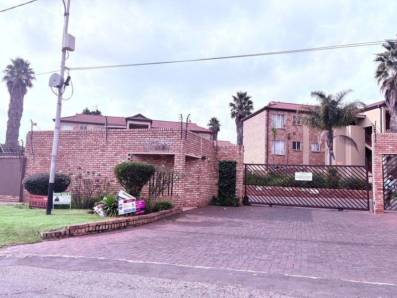 Front entrance view of a brick apartment complex featuring a paved driveway, secure metal gate, and perimeter electric fencing.