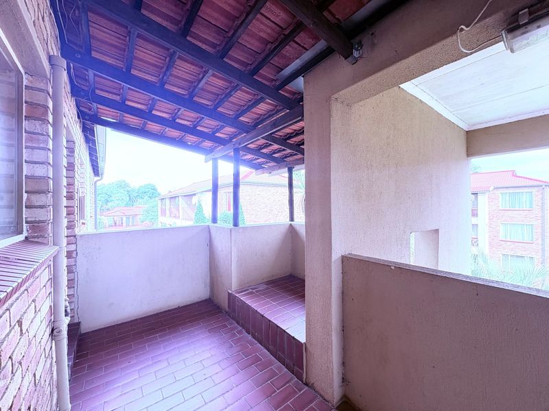A covered balcony featuring tiled flooring and an exposed timber roof structure, offering an outdoor space with views of the surrounding apartment complex.