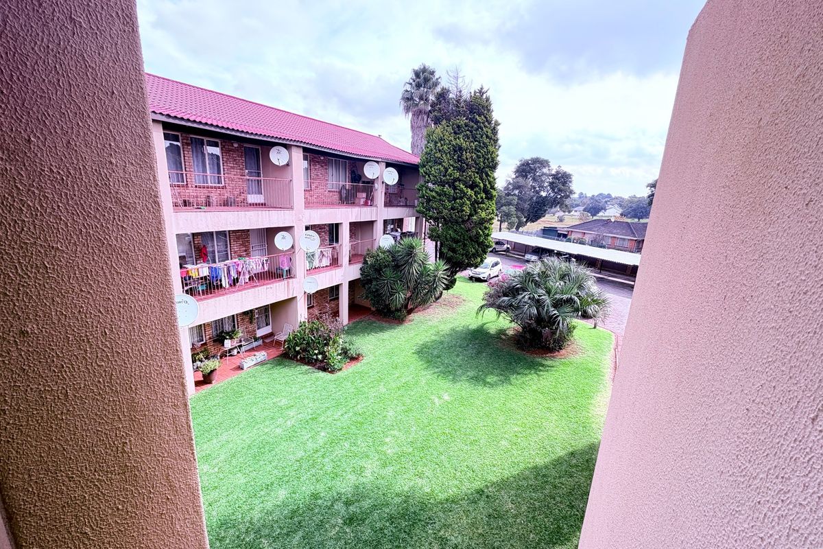 View of the apartment complex rear facade, featuring multi-level balconies overlooking a well-maintained communal garden and covered parking area.